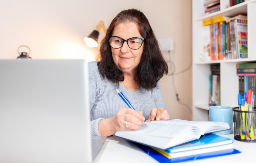 Mulher idosa estudando no computador e fazendo anotações em um livro. O que é encceja.