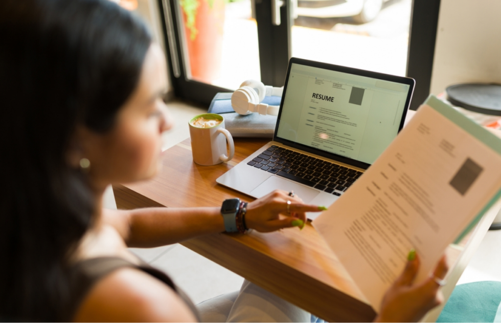 Mulher segurando um currículo na mão em frente a um computador. como colocar graduação no currículo.