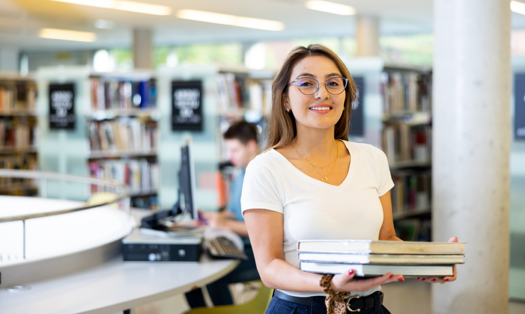 Mulher jovem em biblioteca carregando livros. Valor da faculdade de Administração em diferentes regiões e modalidades.