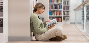 Mulher sentada no chão de uma biblioteca lendo um livro.