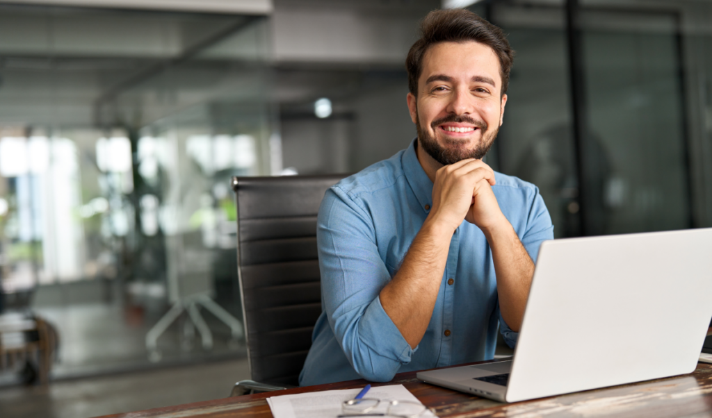 Homem em escritório em frente ao computador sorri para a câmera. Diferença entre especialização e pós-graduação.