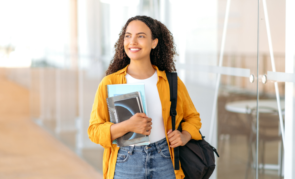 Jovem mulher sorrindo e caminhando. Ela segura alguns cadernos na mão e carrega uma mochila. Prova do eja. 