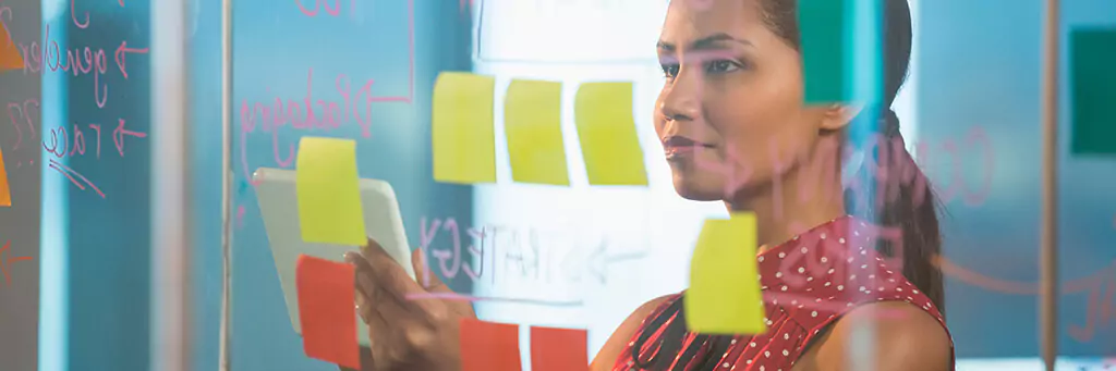 jovem analisando post-it em parede de vidro e segurando tablet