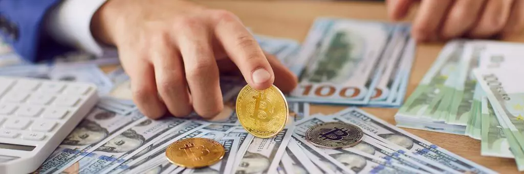 homem segurando com dedo modelo de criptomoeda em cima de mesa com cédulas e calculadora