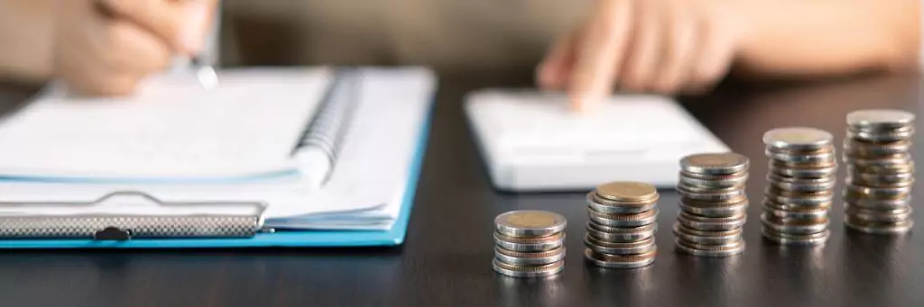pessoa fazendo orçamento com calculadora e caderno, e pilhas de moedas à frente na mesa