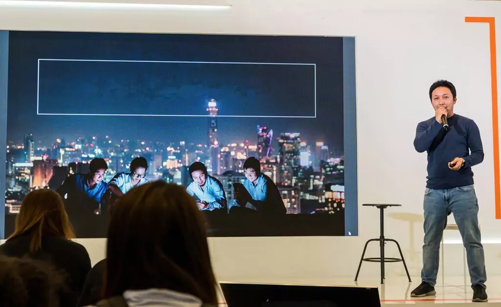 homem dando palestra em palco, com foto projetada na parede e pessoas na plateia