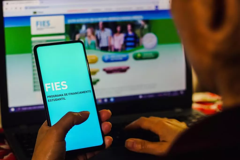 moça olhando app do fies em celular e site do programa ao fundo