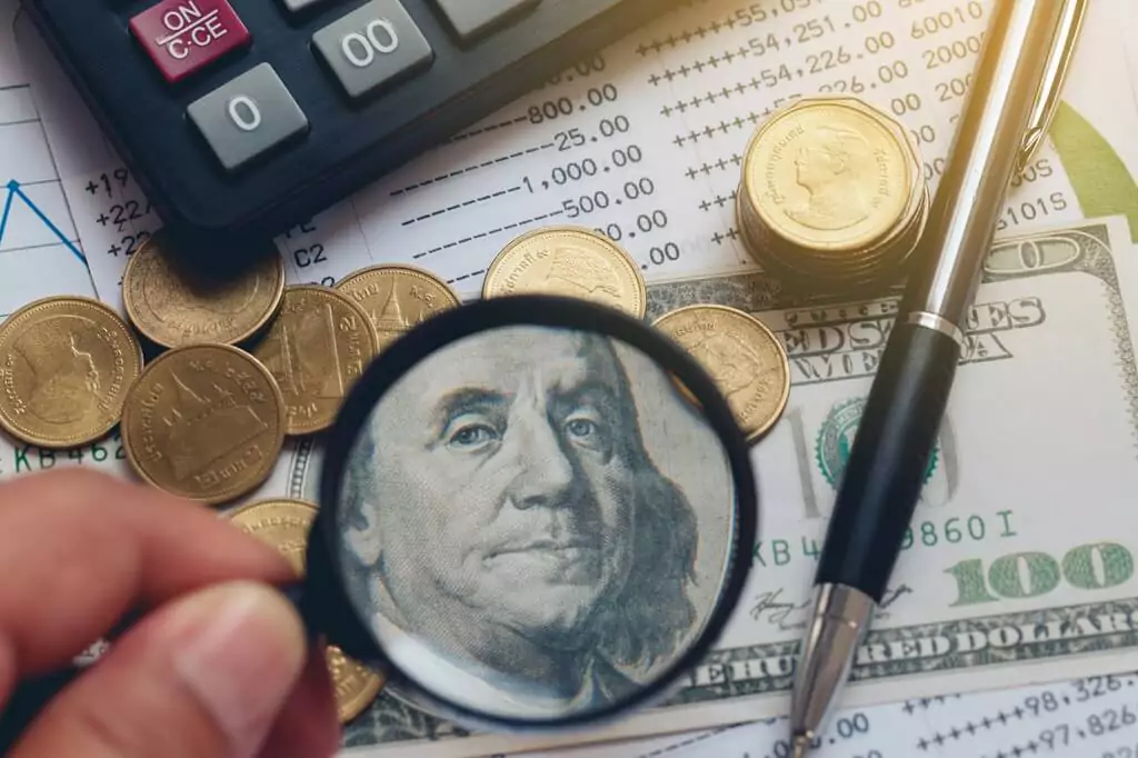 lupa em cima de dólar com moedas, caneta e calculadora ao lado
