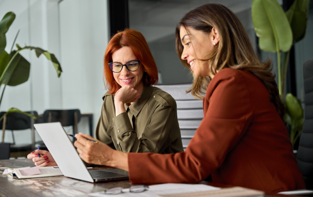 Duas colegas de trabalhando conversando em frente a um computador. Comunicação assertiva.