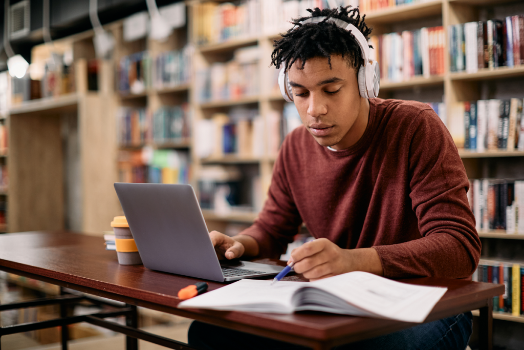 homem lendo livro em frente ao computador na biblioteca. Curso de história na Anhanguera.