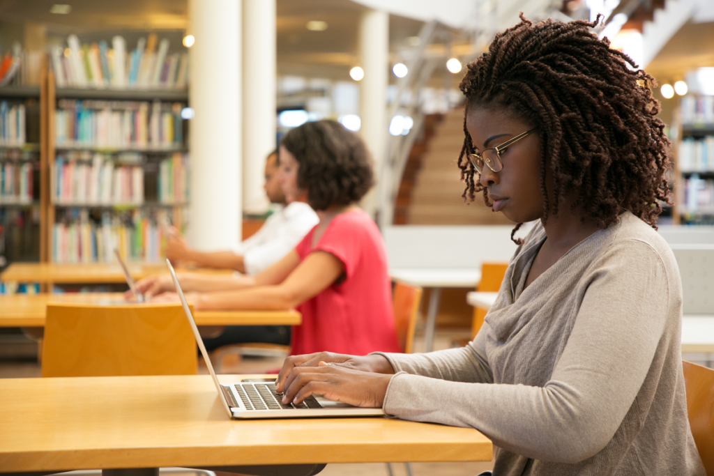 Mulher escrevendo no computador em biblioteca. Vale a pena investir no curso de letras português e inglês.